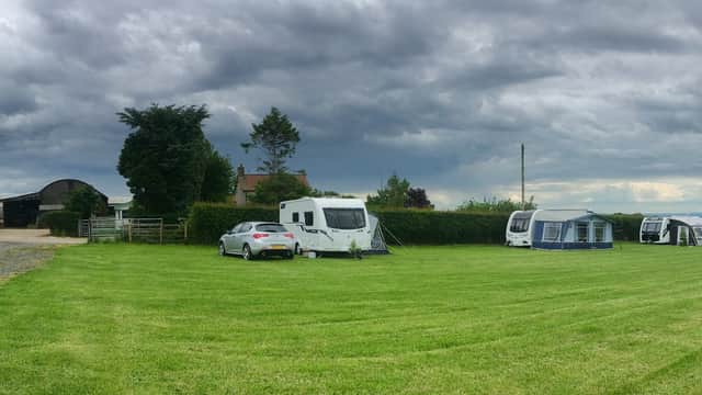 Caravans and tents set in a grassy field under a cloudy sky, with trees and farm buildings in the background.
