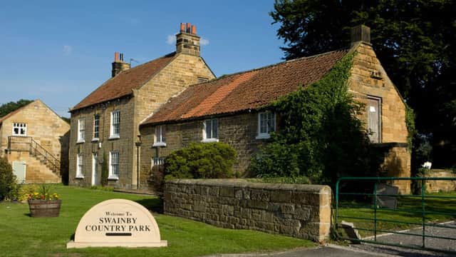 Stone buildings with red roofs, green grass, and a welcoming sign for Swainby Country Park in a sunny landscape.