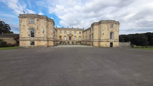 Gilling Castle with stone facade and large central entrance, surrounded by a paved area and lush greenery in the background.