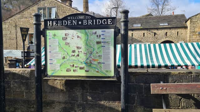 Welcome sign with a map of Hebden Bridge, stone buildings, and striped market tents in a park setting.