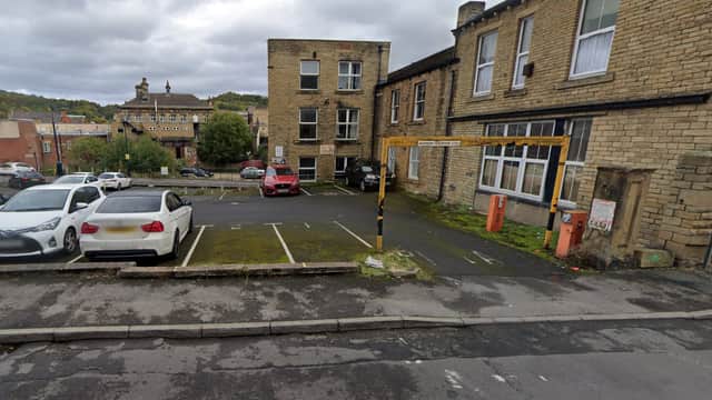 Car park with several vehicles, stone buildings in the background, and a cloudy sky in Dewsbury, Yorkshire.