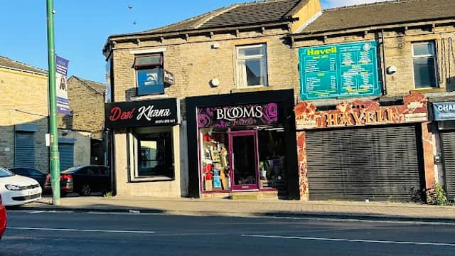 Bright storefront of "Blooms of Bradford" with large windows, colorful signage, and a clear blue sky above.