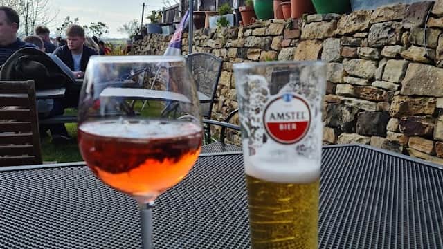 A glass of rosΓ© wine and a pint of Amstel beer on a patio table, with a stone wall and potted plants in the background.