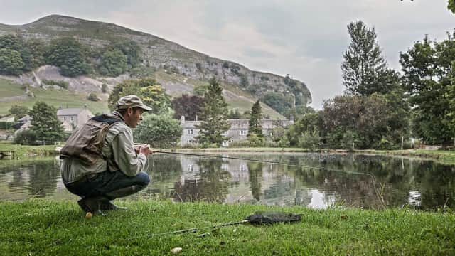 A man crouches by a pond, fishing, with lush greenery and limestone hills in the background.
