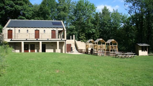 Modern building with wooden doors, surrounded by green grass and trees, featuring picnic tables and wooden structures nearby.
