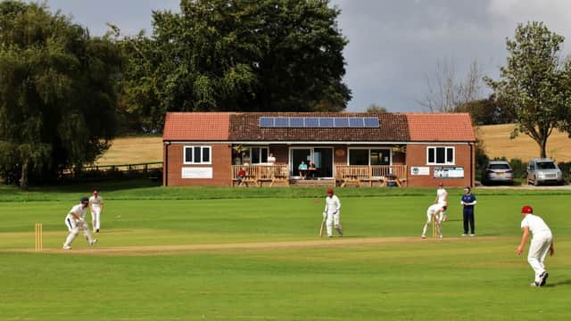 Cricket match in progress on a green field with a club house featuring solar panels in the background.