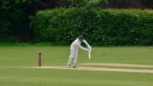 Cricketer in white uniform batting on a green field with a red cricket ball and stumps in the background.