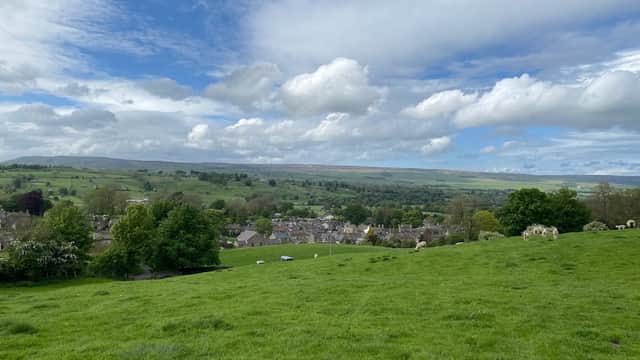 Lush green hills and fields overlook a quaint village under a bright blue sky with fluffy white clouds.