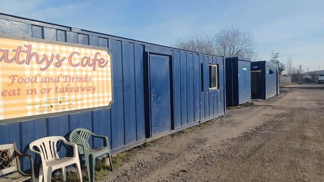 Blue shipping containers line a gravel path, with chairs in front of a cafe sign offering food and drinks.