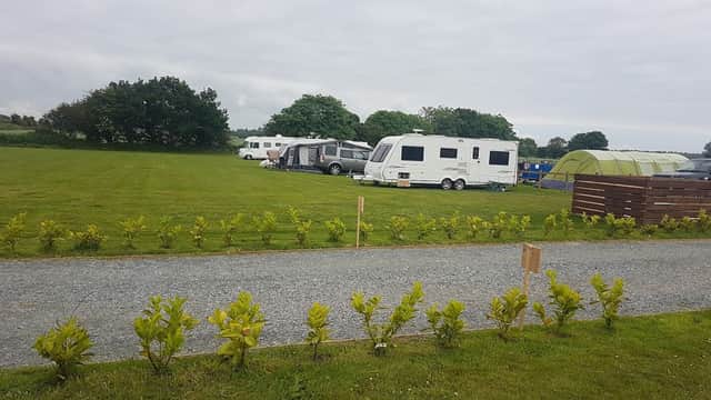 Caravans parked on a grassy field, bordered by a neat hedge, with trees and camping tents in the background.