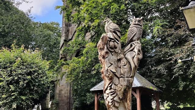 Car park with pay & display signs, surrounded by trees and a carved wooden sculpture near a church tower.