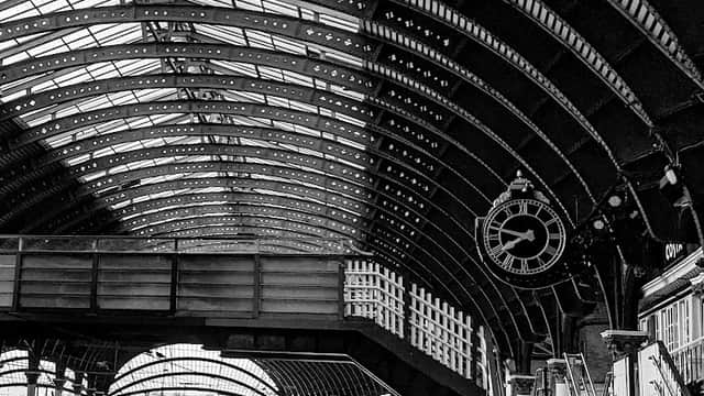 Historic train station interior with arched metal roof, clock, and stairway leading to platforms. Black and white photo.