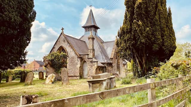 Historic stone church with a tall spire, surrounded by trees and a grassy cemetery, near a wooden fence.