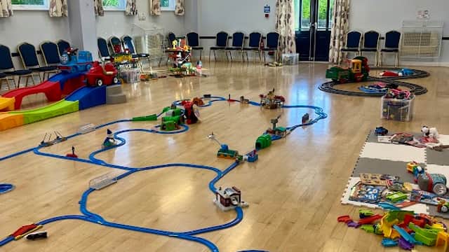 Bright, spacious village hall with train tracks, toys, and colorful seating, featuring large windows and a stained glass window.