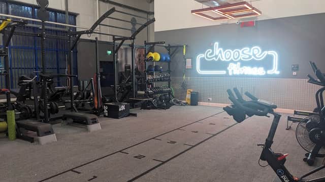 Bright gym interior with workout equipment, weights, and a neon sign reading "choose Fitness."