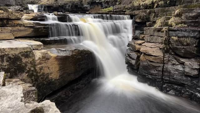 A cascading waterfall flows over rocky ledges in a serene landscape surrounded by trees and mossy stones.