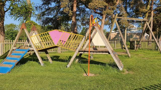 Colorful playground equipment with swings and a climbing frame surrounded by grass and trees in a fenced area.