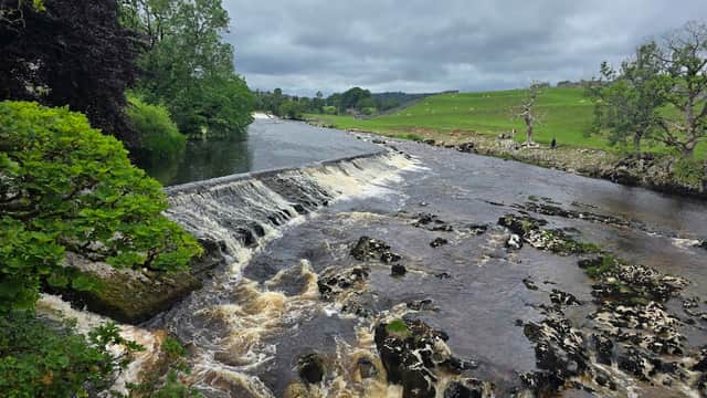 A scenic view of a river waterfall with lush greenery and rolling hills under a cloudy sky in the Yorkshire Dales.