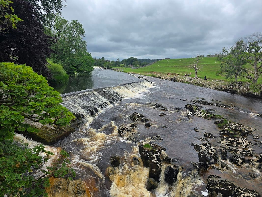 A scenic view of a river waterfall with lush greenery and rolling hills under a cloudy sky in the Yorkshire Dales.