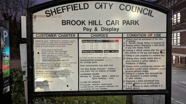 Sign for Brook Hill Car Park detailing customer charges, conditions, and operational hours, set against a dimly lit background.