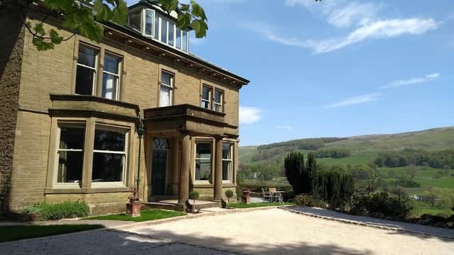 Victorian-style house with large windows, surrounded by greenery and hills under a clear blue sky.