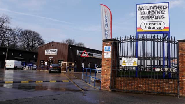 Milford Building Supplies entrance with a sign, vehicles in the lot, and trees in the background. Wet pavement visible.