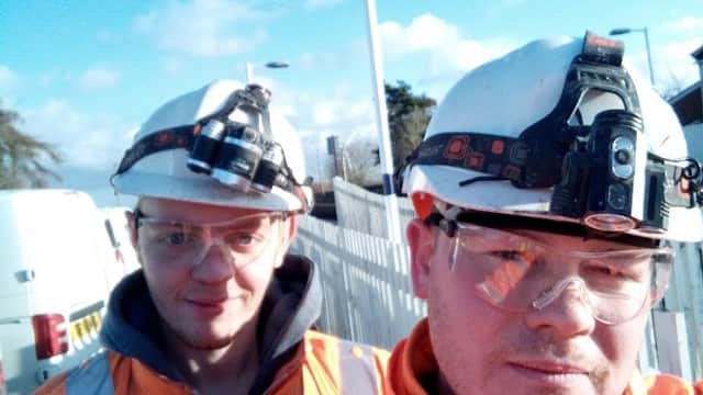 Two workers in orange safety jackets and helmets with headlamps, standing near a bus stop under a clear blue sky.